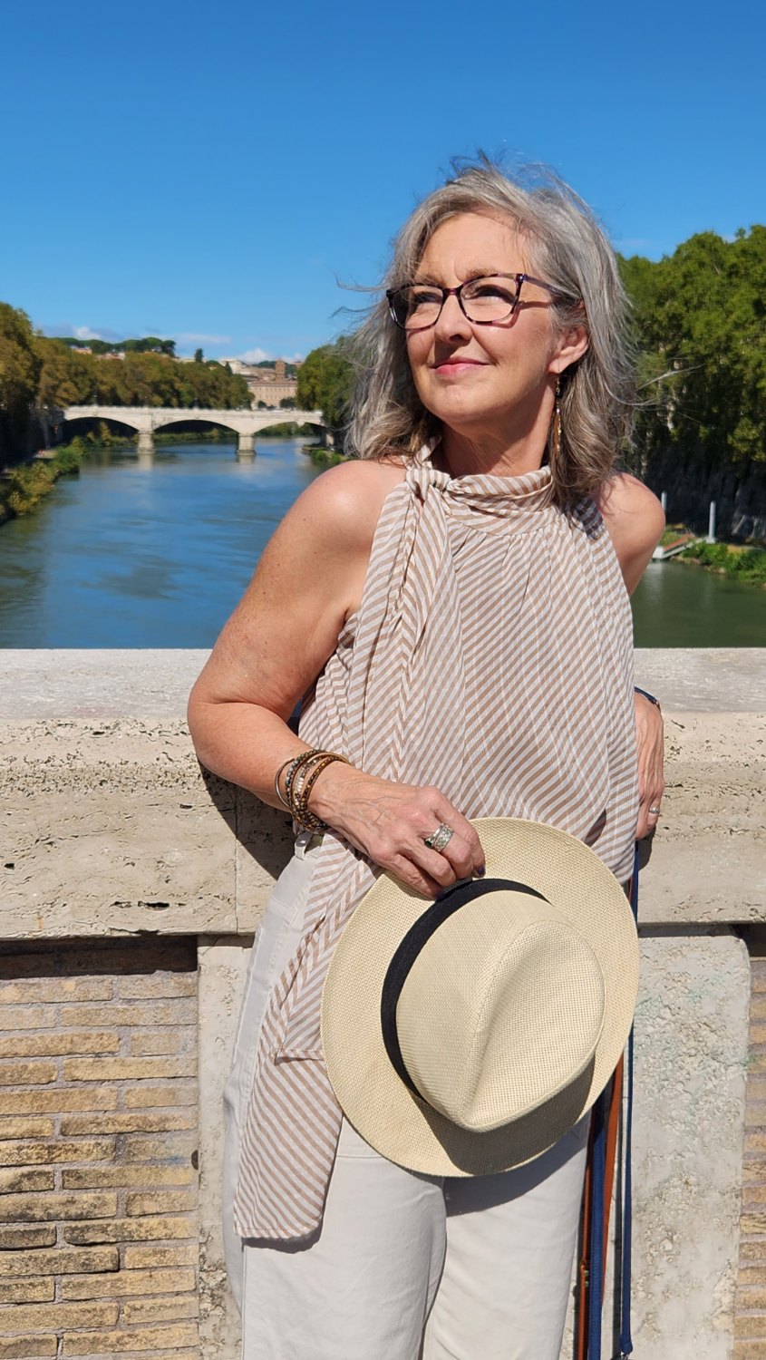 Gina Calvert smiling by the river in Rome, holding a straw hat